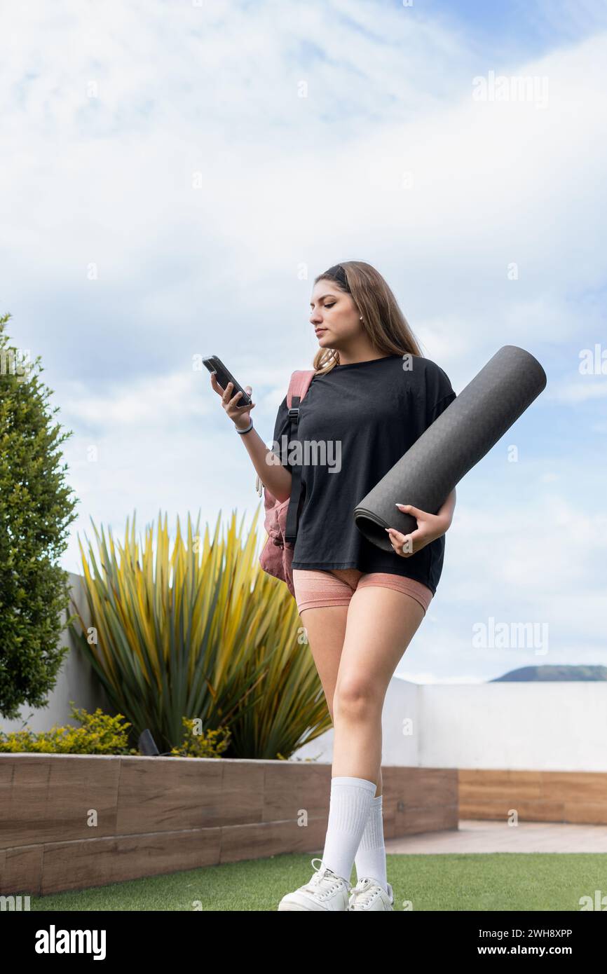Jeune femme latina marchant à la salle de gym le matin avec son sac à dos et tapis regardant le téléphone portable tout en marchant Banque D'Images