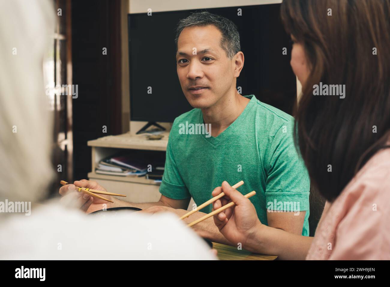 Homme japonais mangeant à la maison avec des baguettes regarde et sourit à sa famille. Banque D'Images
