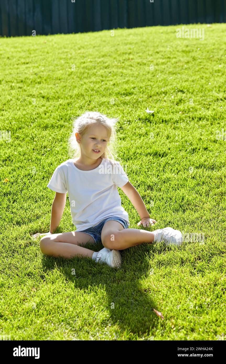 Petite fille assise sur la pelouse dans le parc de la ville d'été. Enfance, loisirs et concept de personnes - repos heureux de l'enfant et passer un bon moment Banque D'Images