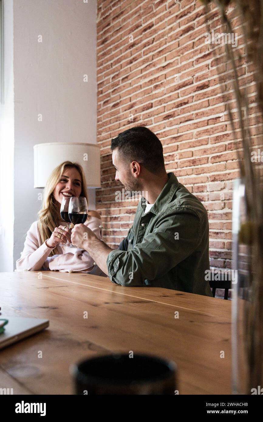 Couple affectueux souriant dans une date buvant de l'alcool assis à table dans une maison. Femme joyeuse et homme buvant du vin ensemble dans un salon avec Banque D'Images
