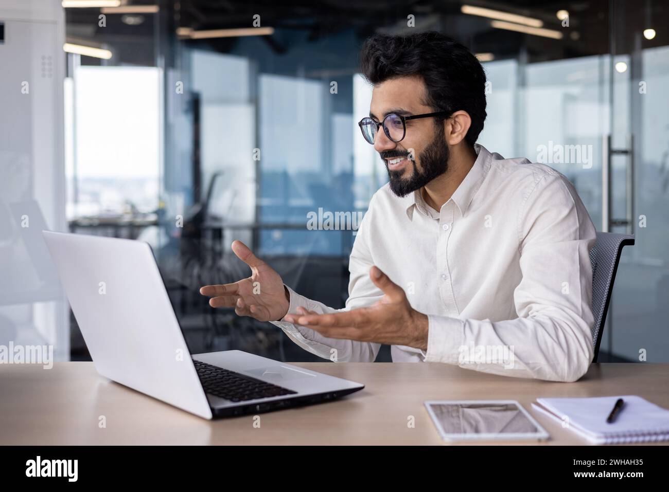 Réunion en ligne, conférence. Jeune homme indien, employé de bureau et homme d'affaires prie sur ordinateur portable pour l'appel vidéo, assis dans le bureau au bureau, faisant des gestes avec les mains et souriant. Banque D'Images