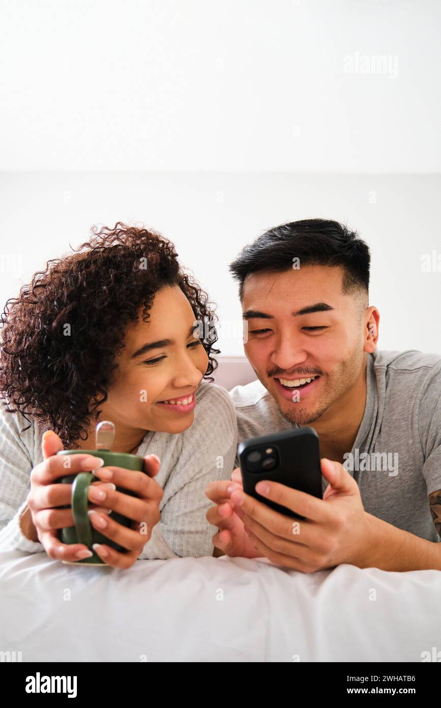 Couple heureux multiethnique utilisant le smartphone sur le lit. Banque D'Images