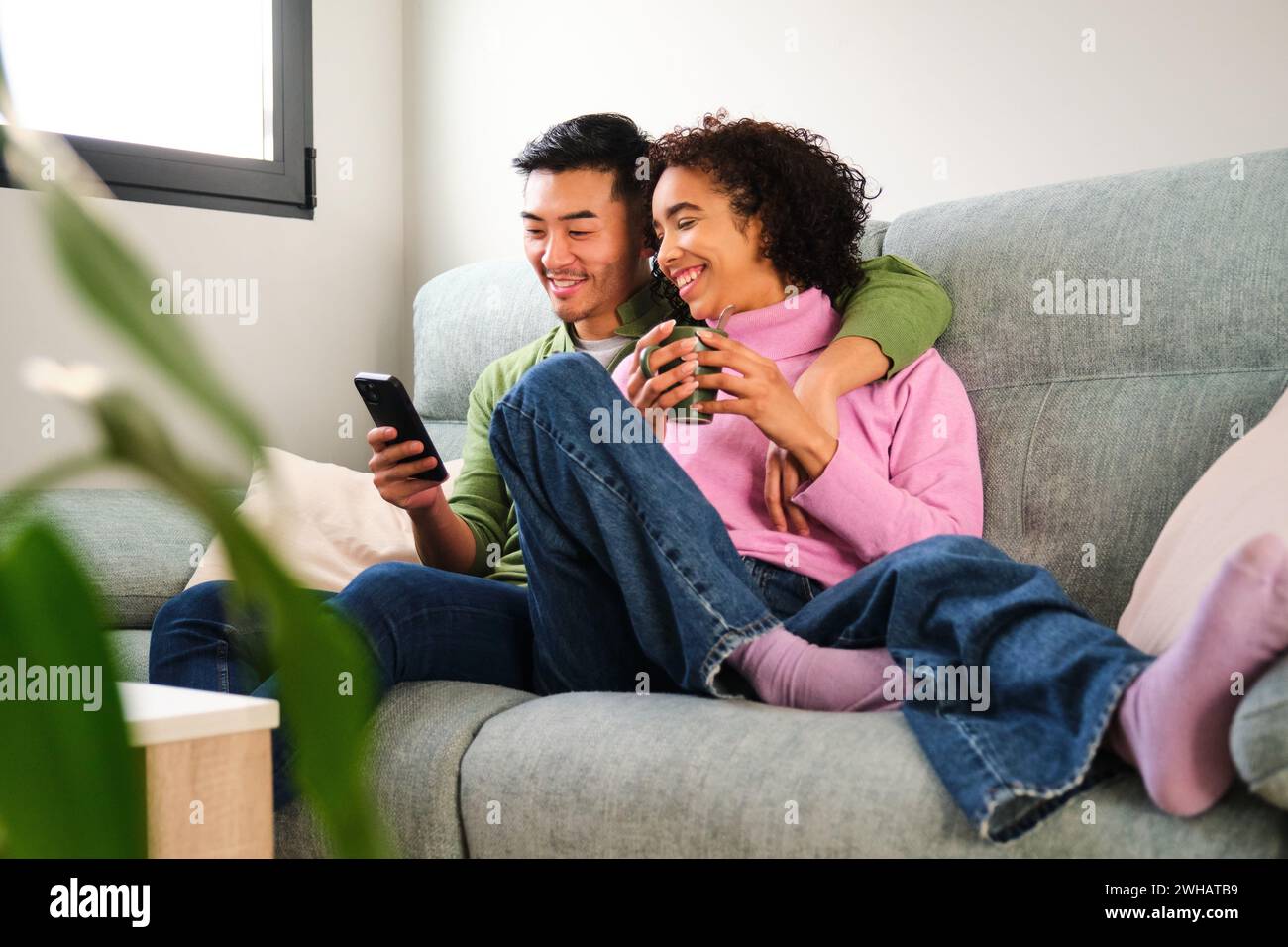Couple heureux multiethnique utilisant le smartphone ensemble sur le canapé. Banque D'Images