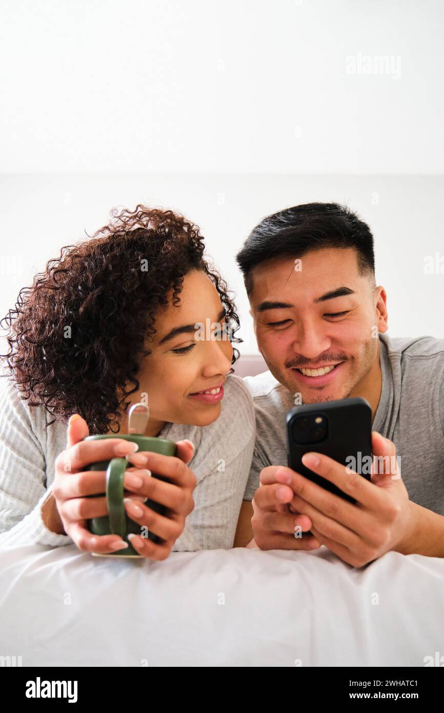 Couple multiracial souriant et utilisant le téléphone sur le lit. Banque D'Images