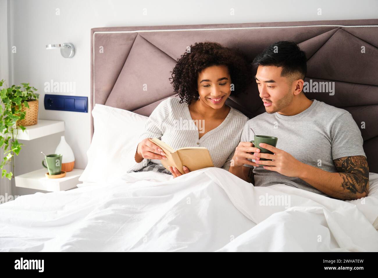 Couple multiethnique souriant et lisant un livre sur le lit ensemble. Banque D'Images