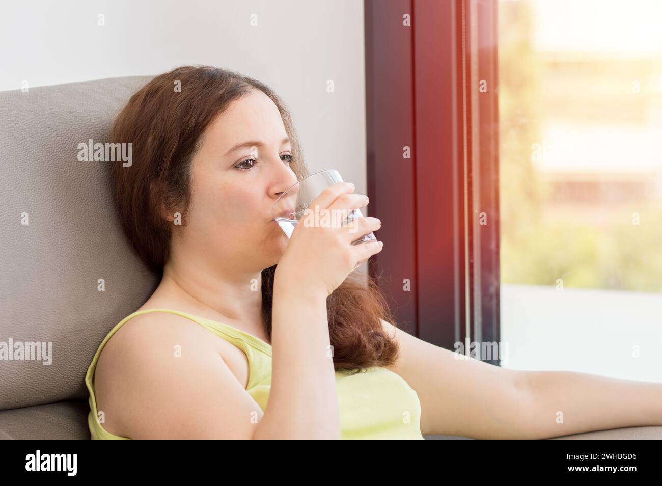 Femme heureuse buvant de l'eau fraîche d'un verre assis sur un canapé dans le salon à la maison avec une fenêtre sur une éruption de soleil Banque D'Images