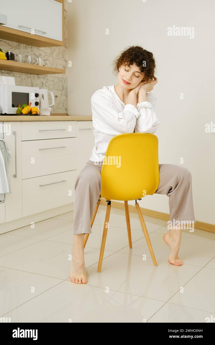 Jeune femme rêveuse assise sur une chaise dans la cuisine les yeux fermés Banque D'Images