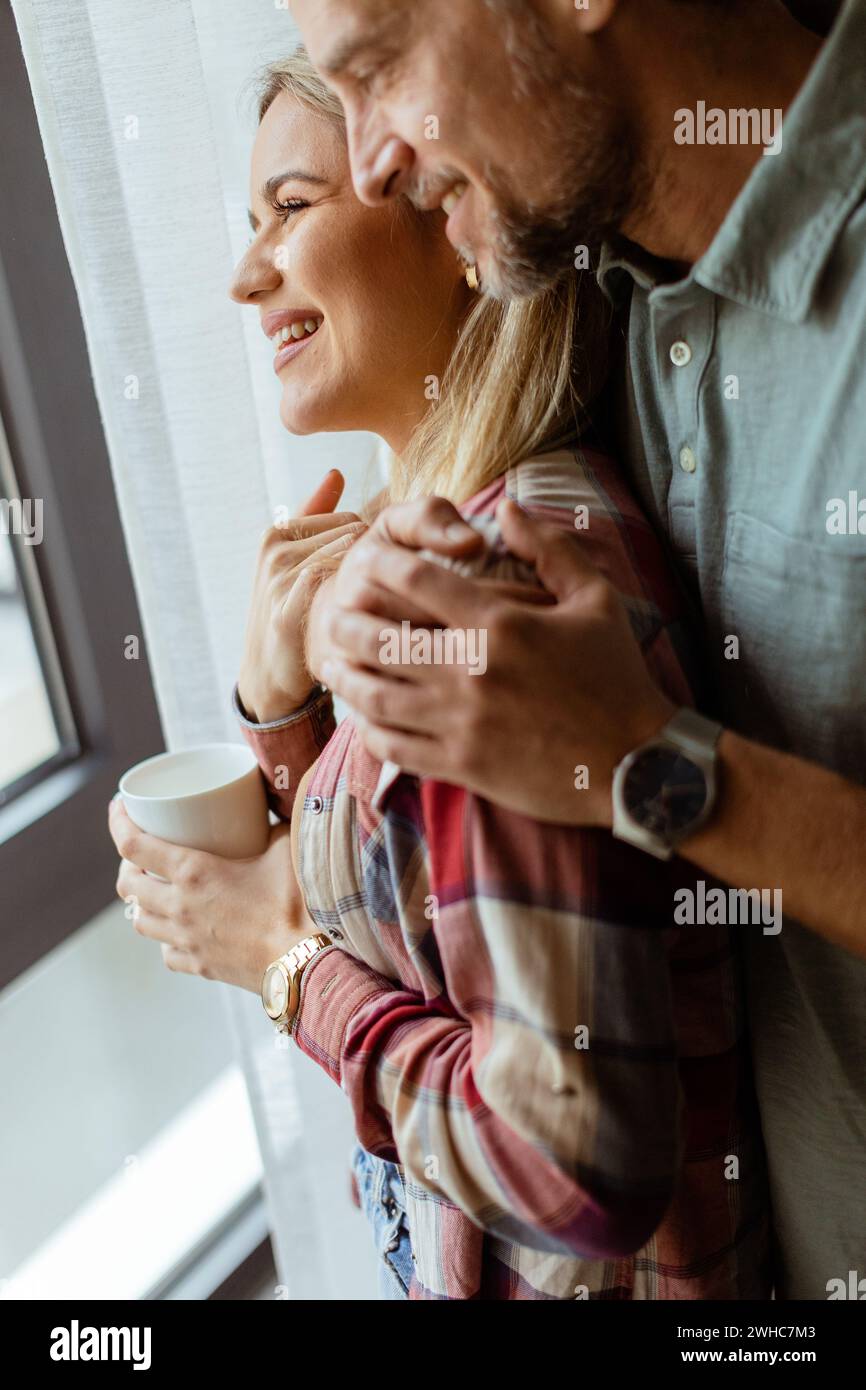 Dans une pièce légèrement éclairée, un homme embrasse amoureusement une femme par derrière alors qu'ils regardent tous les deux par la fenêtre, partageant ensemble un moment calme et intime Banque D'Images