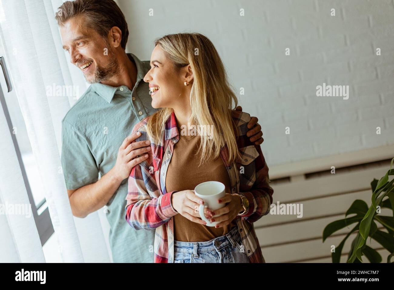 Dans une pièce légèrement éclairée, un homme embrasse amoureusement une femme par derrière alors qu'ils regardent tous les deux par la fenêtre, partageant ensemble un moment calme et intime Banque D'Images