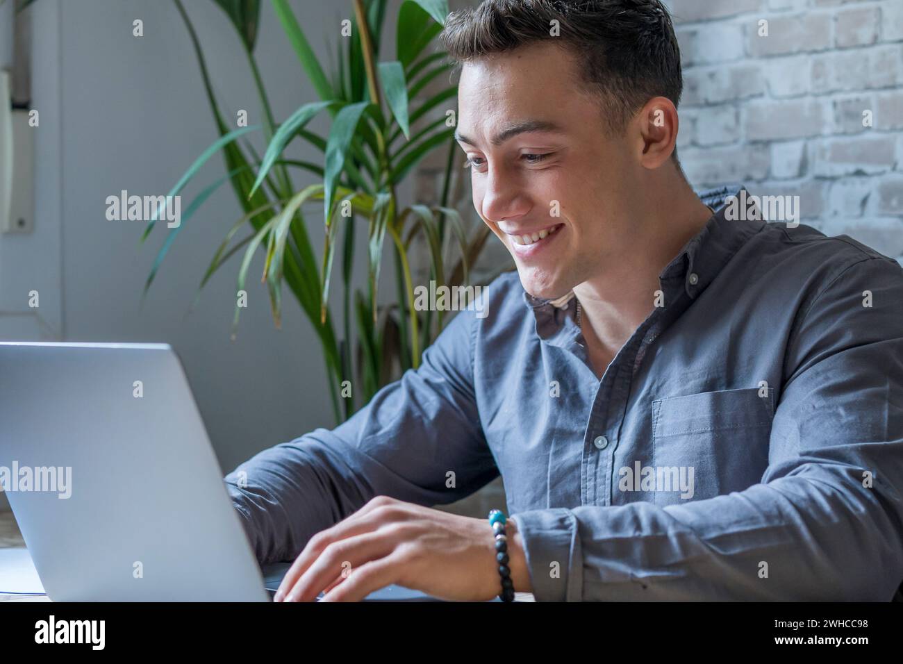 Vue latérale beau jeune homme d'affaires dans les lunettes travaillant avec l'ordinateur à distance, assis à la table en bois dans le bureau. Homme heureux agréable communiquant dans le réseau social, recherche d'informations en ligne. Banque D'Images