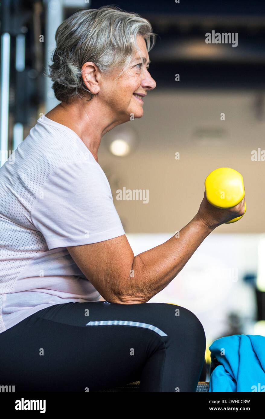 femme mature à la salle de gym formant son corps et ses biceps assis dans un banc avec un haltère dans sa main - mode de vie senior sain et fitness Banque D'Images