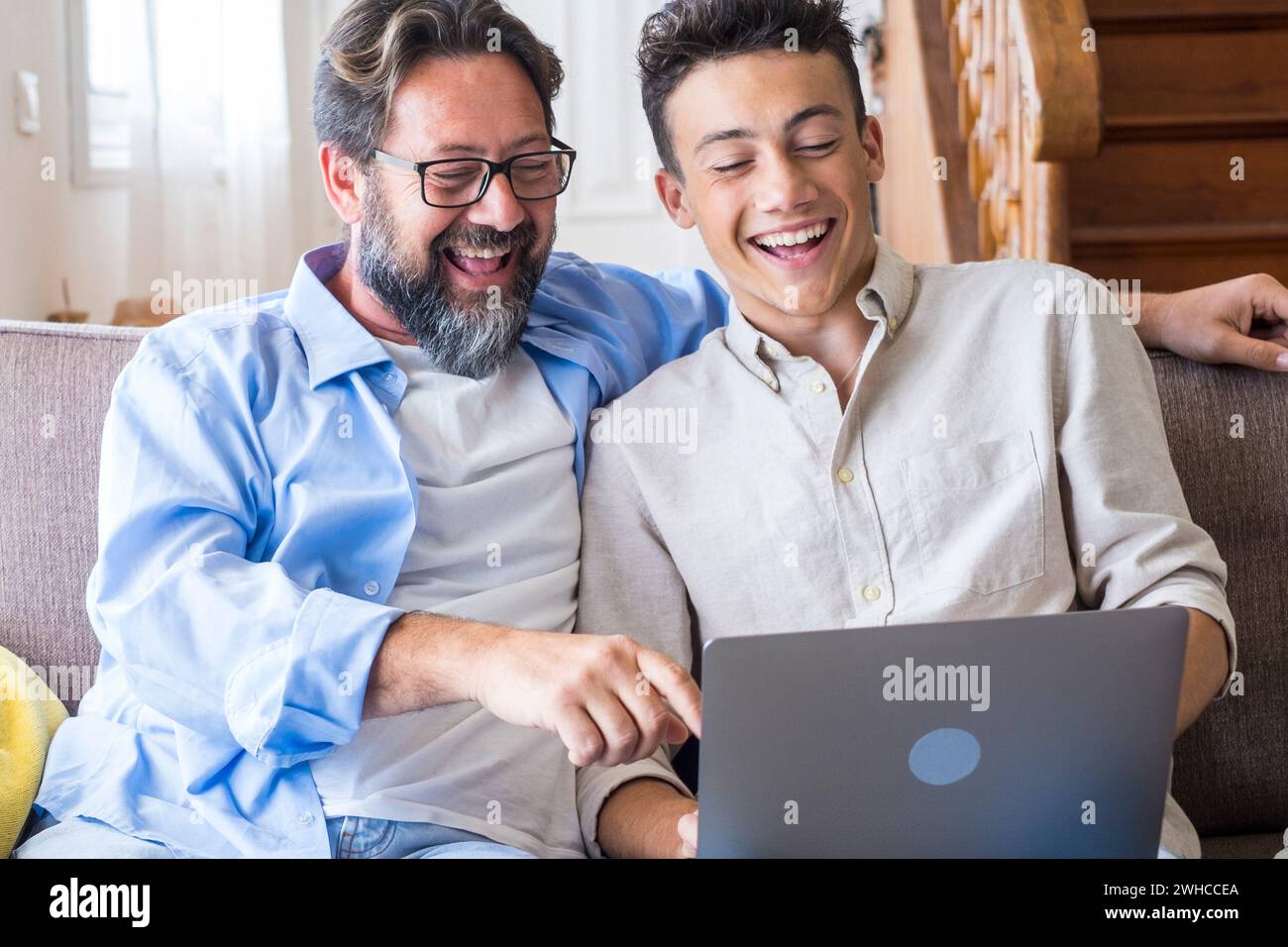 Heureux jeune père assis sur le canapé à l'aide d'un ordinateur portable se détendre avec le fils étudiant tenant smartphone amusez-vous ensemble, papa souriant et petit garçon adolescent profiter du week-end à la maison repos sur le canapé occupé avec des gadgets Banque D'Images