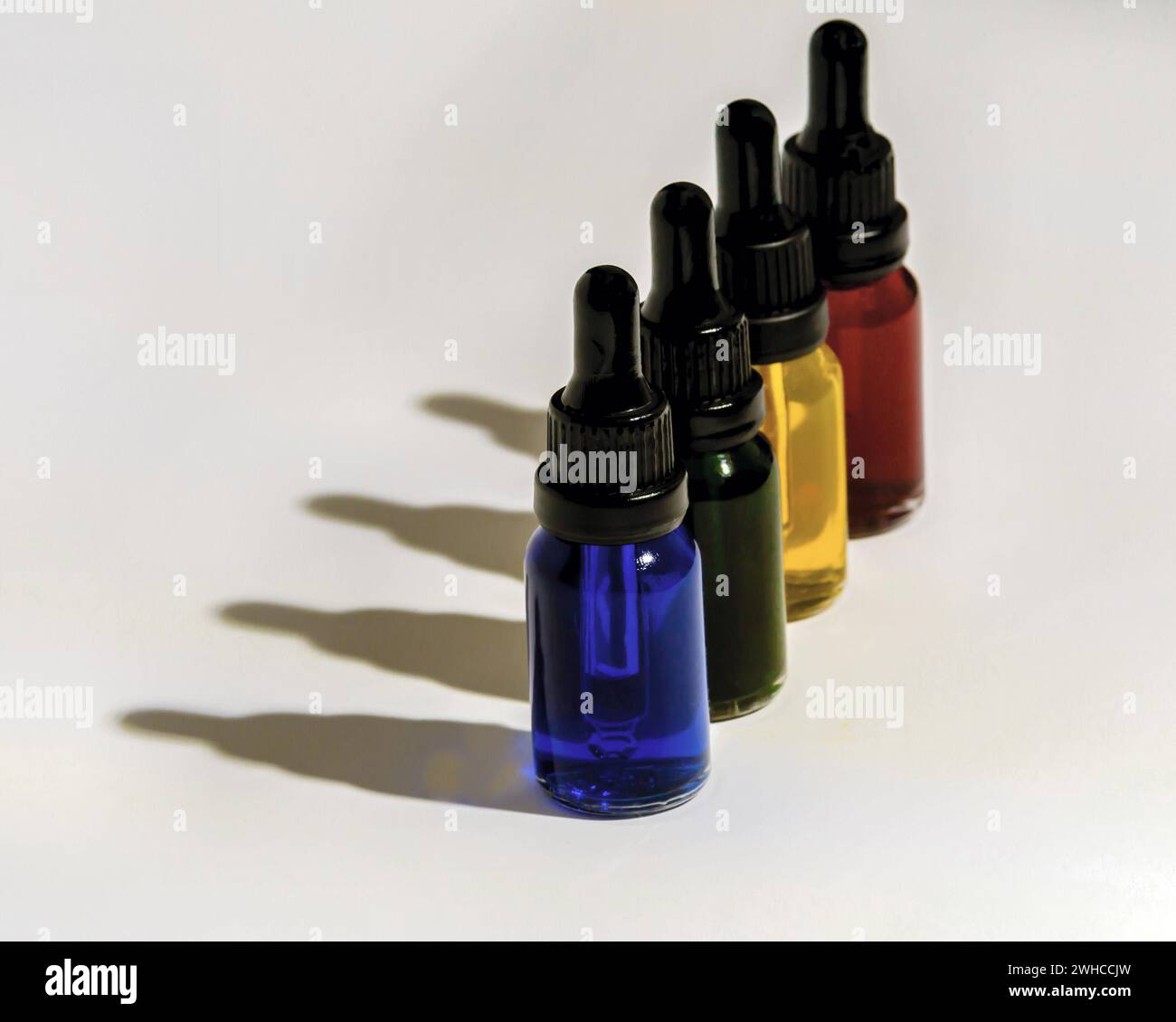 Quatre flacons compte-gouttes en verre remplis de liquide bleu, vert, jaune et rouge projetant des ombres sombres sur fond blanc Banque D'Images