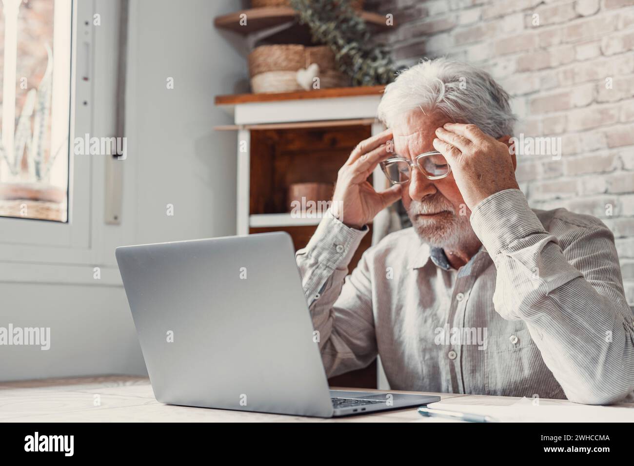 Homme fatigué mature des années 70 travaillant à l'ordinateur portable de la maison, enlevant les lunettes, touchant la tête et les yeux. Vieil homme retraité souffrant de maux de tête, vertiges après avoir regardé l'écran d'ordinateur. Problème de vision Banque D'Images