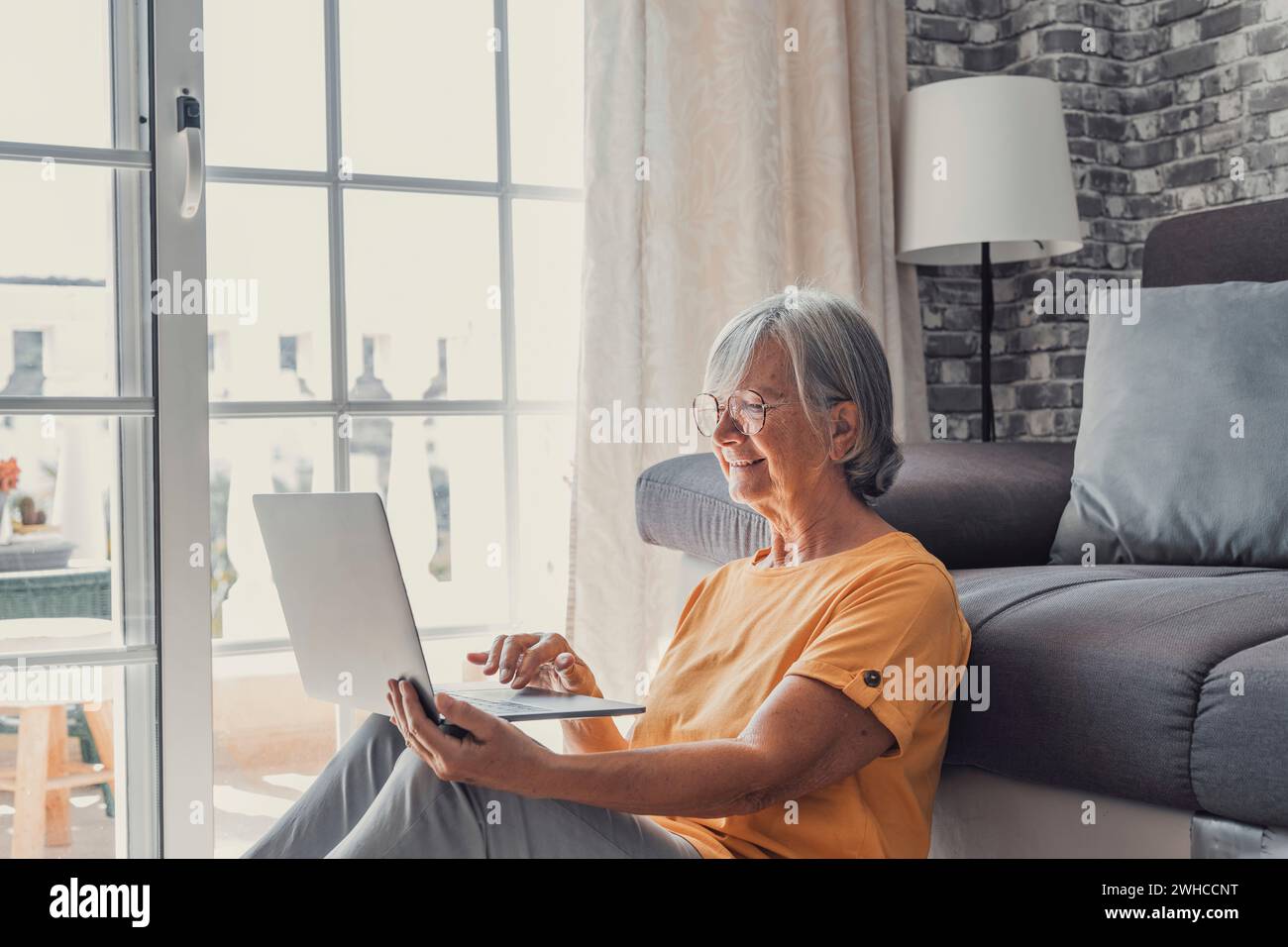 Souriante femme caucasienne d'âge moyen assis à côté du canapé dans le salon naviguant sur Internet sans fil sur l'ordinateur, femme âgée moderne heureuse se détendre sur le sol à la maison en utilisant un appareil pc portable, concept de technologie âgée Banque D'Images