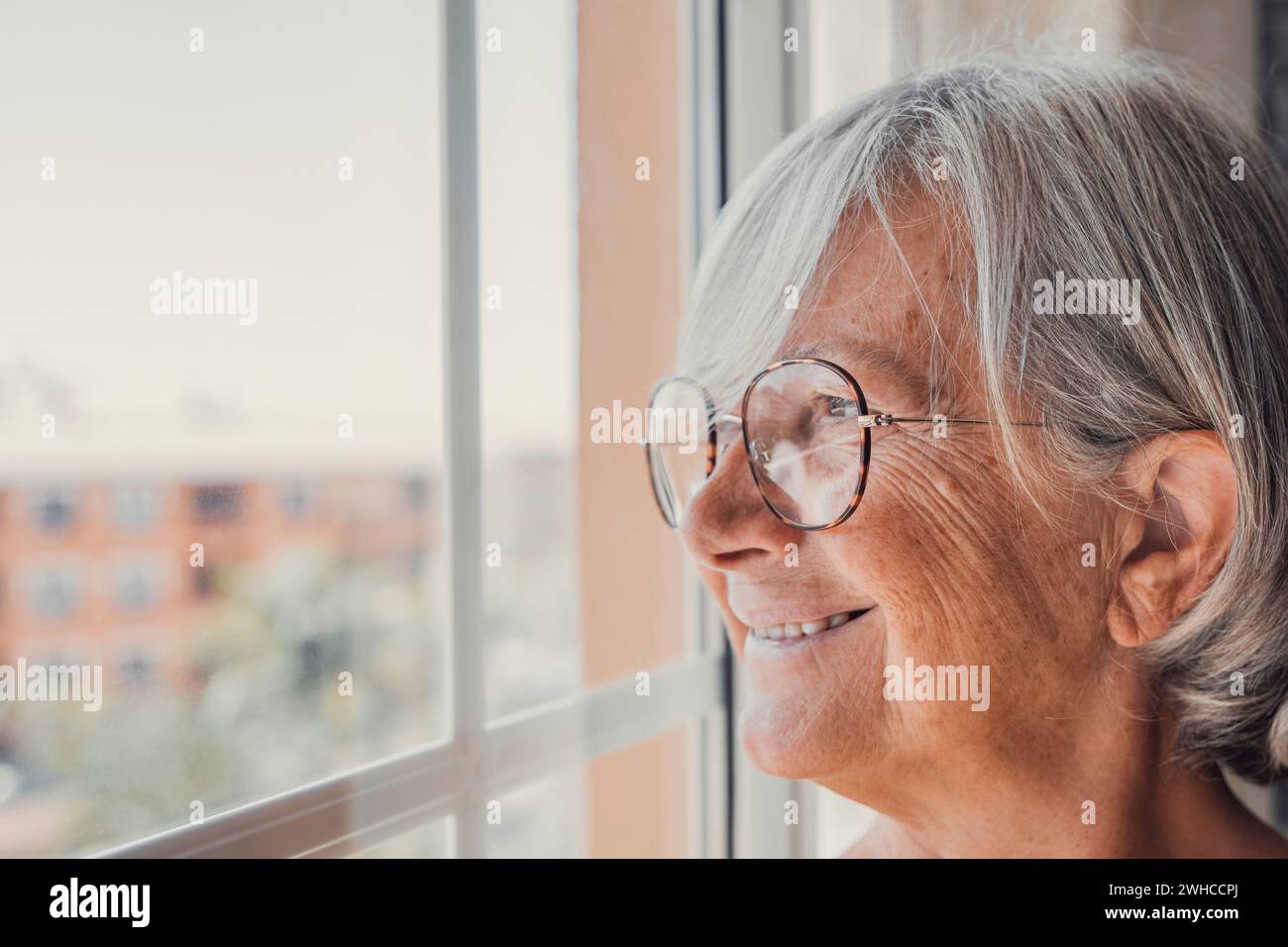 Rêveur âgée souriante des années 60 Hoary femme regardant à distance hors de la fenêtre, se rappelant de bons souvenirs ou visualisant l'avenir à la maison, la planification de vacances ou profiter d'une journée paisible consciente seul à l'intérieur. Banque D'Images