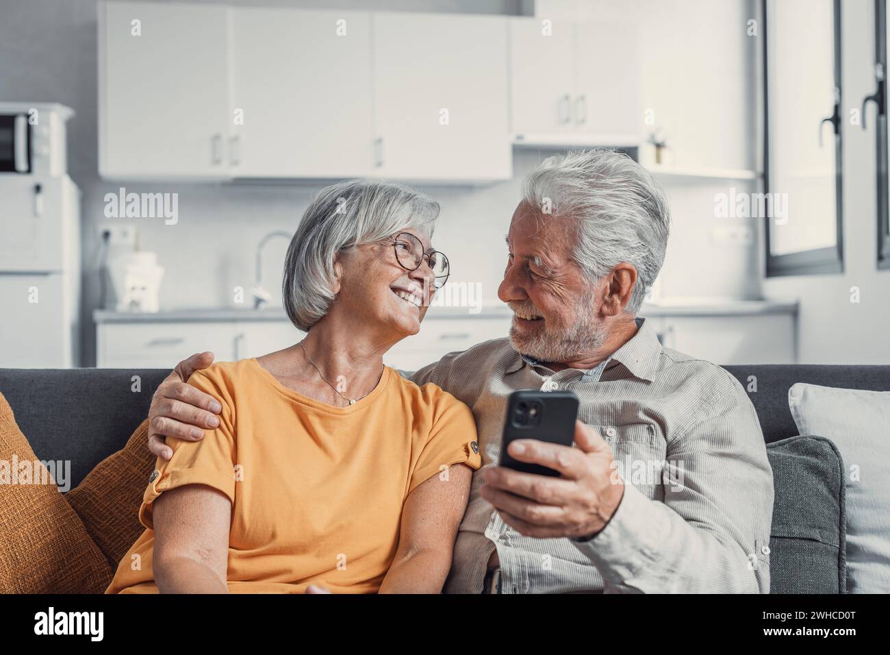Heureuse famille mature, femme et mari utilisant le téléphone ensemble à la maison, souriant homme et femme d'âge moyen assis sur le canapé, en utilisant des applications pour appareils mobiles, regardant des vidéos dans le réseau social, surfant sur Internet Banque D'Images