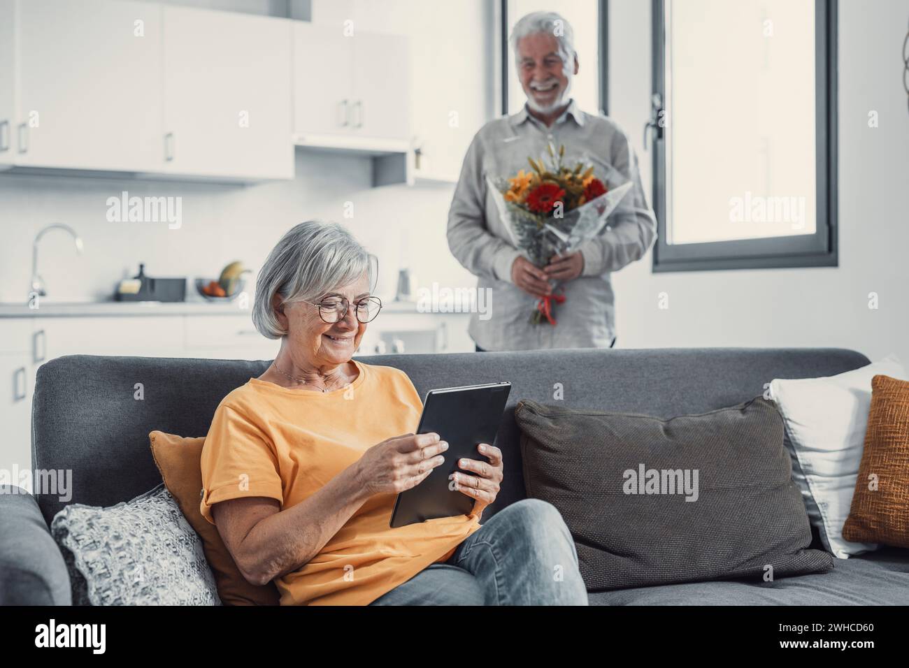 Vieil homme donnant des fleurs à sa femme assise sur le canapé à la maison pour le jour de San Valentinesí. Les retraités apprécient la surprise ensemble. Dans l'amour les gens s'amusent. Banque D'Images