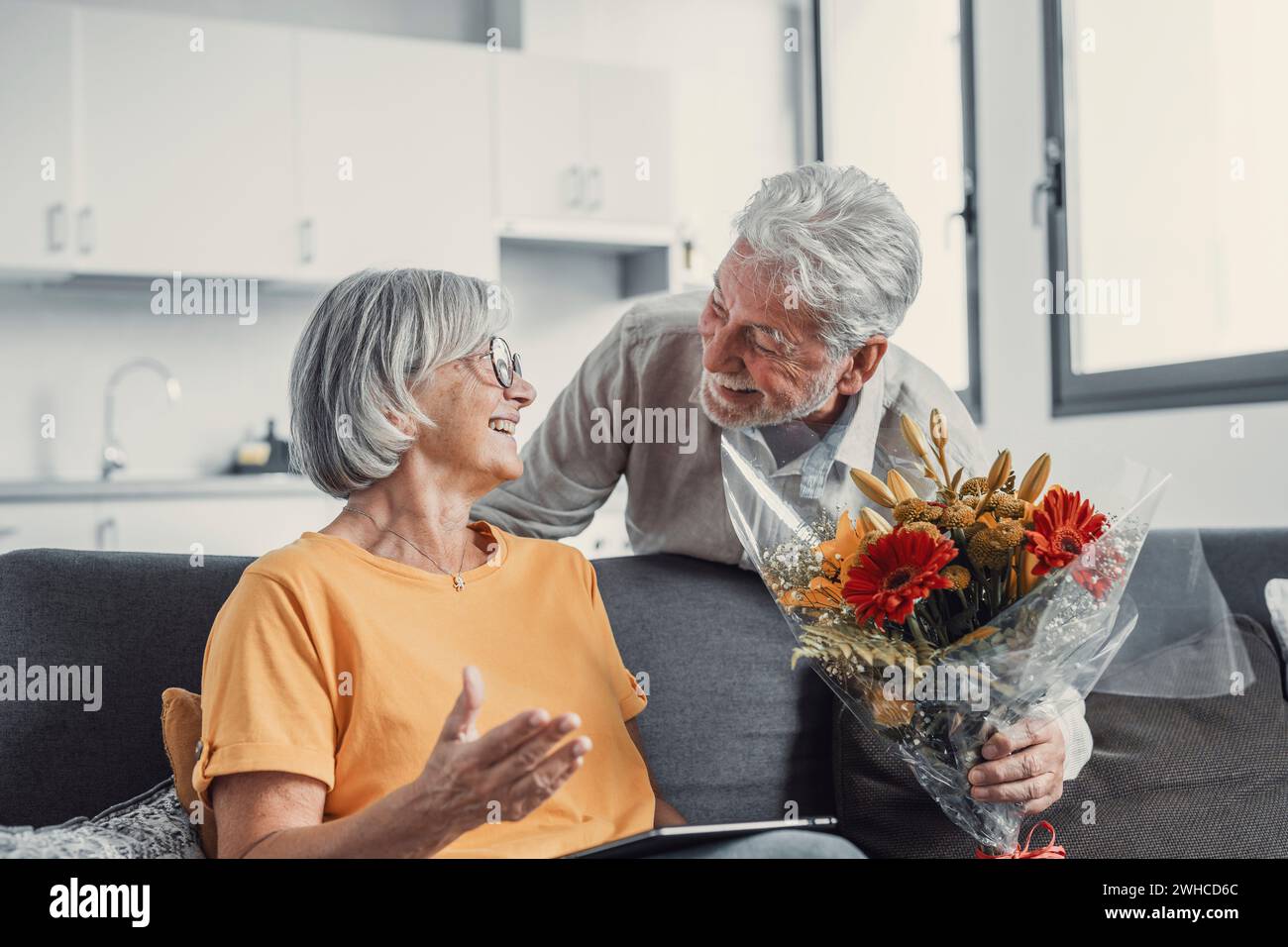 Vieil homme donnant des fleurs à sa femme assise sur le canapé à la maison pour le jour de San Valentinesí. Les retraités apprécient la surprise ensemble. Dans les gens amoureux havi Banque D'Images