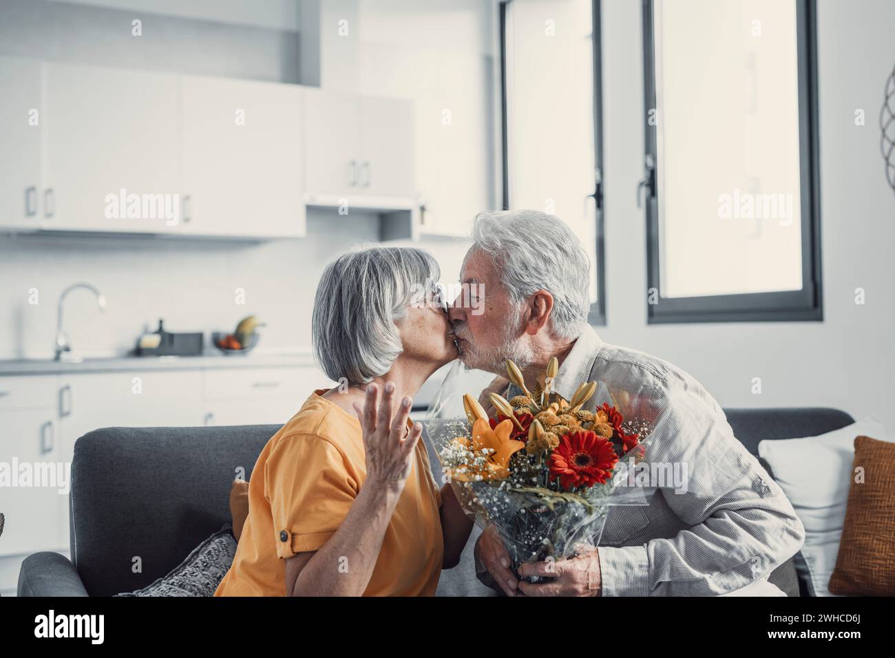 Vieil homme donnant des fleurs à sa femme assise sur le canapé à la maison pour le jour de San Valentinesí. Les retraités apprécient la surprise ensemble. Dans l'amour les gens s'amusent. Banque D'Images