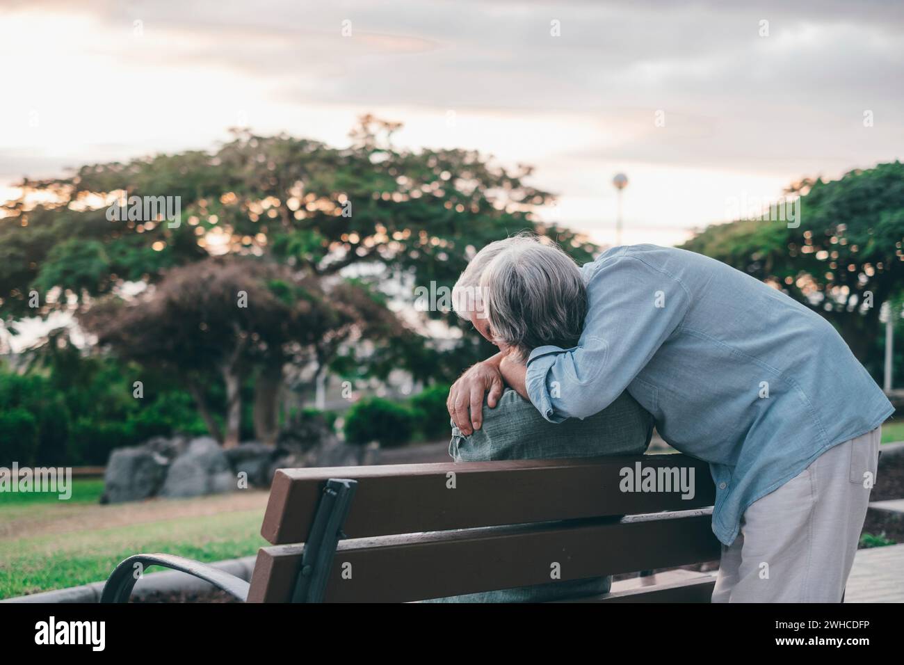 Femme d'âge mûr assise sur le banc du parc avec un vieil homme l'embrassant par le dos. Couple de personnes retraitées se relaxant et profitant du coucher de soleil ensemble à l'extérieur. Banque D'Images