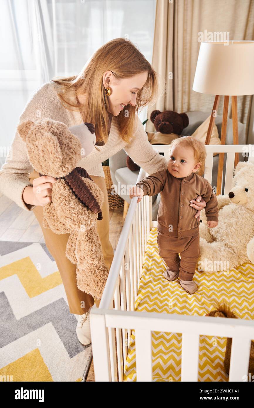femme tenant l'ours en peluche près du petit fils mignon debout dans la crèche dans la chambre de bébé, maternité heureuse Banque D'Images