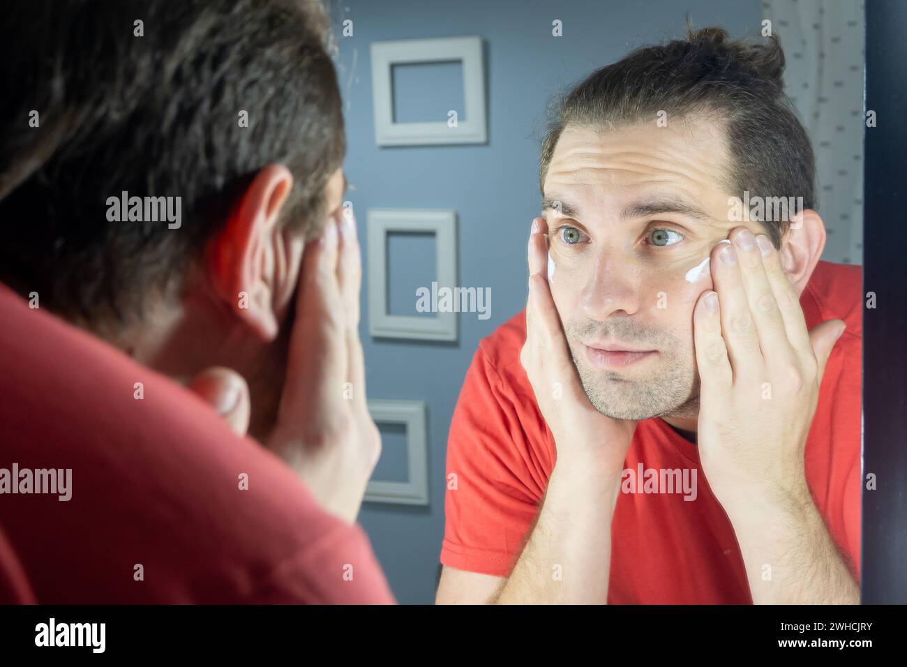 Homme de soins de la peau. L'homme applique de la crème sur son visage devant le miroir. Soins quotidiens de la peau pour un homme. Photo naturelle à la maison. Banque D'Images