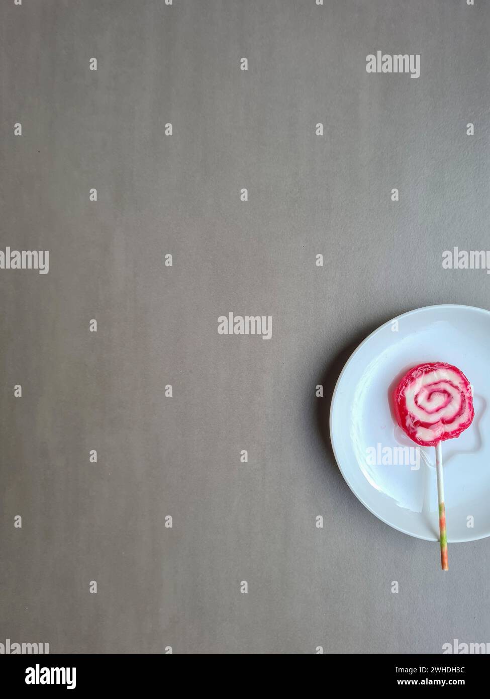 Une sucette rouge et blanche avec un bâton repose sur une plaque sur un fond gris clair Banque D'Images