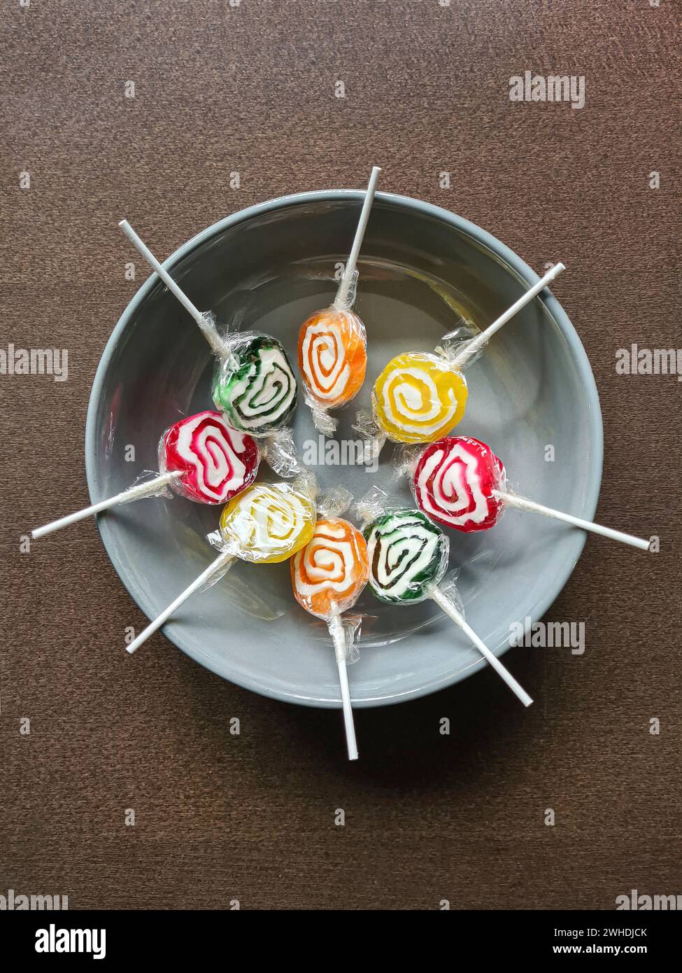 Huit sucettes colorées sont encore enveloppées dans une feuille de plastique en cercle dans un bol gris Banque D'Images