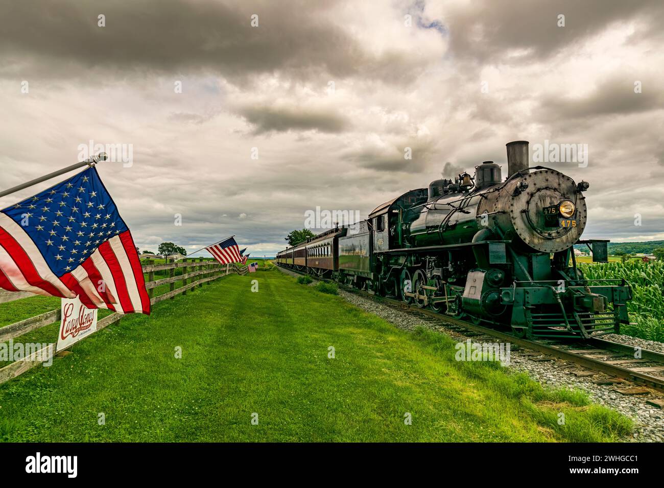 Les drapeaux américains agitent dans le vent, sur une clôture, tandis qu'un train de passagers à vapeur approche Banque D'Images