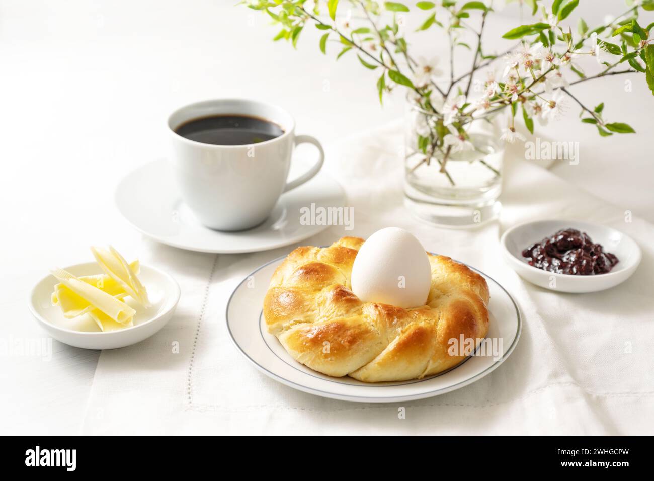 Pain de levure de Pâques tressé en forme de nid avec un œuf, du café, du beurre et de la confiture comme petit déjeuner festif du dimanche sur une table blanche, copie Banque D'Images