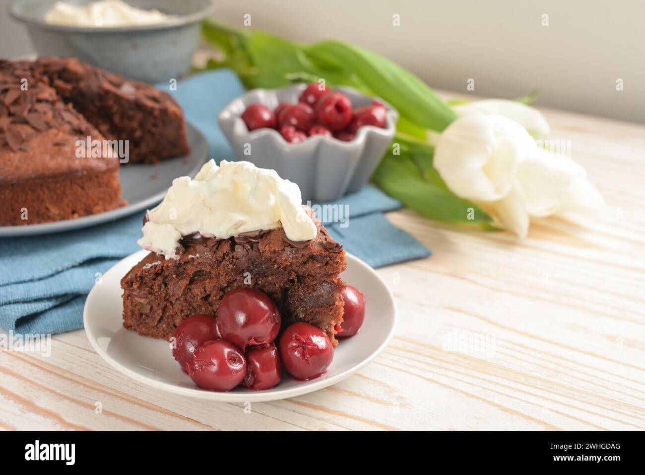 Gâteau au chocolat maison juteux avec cerises morello et crème fouettée, quelques fleurs de tulipe et une serviette bleue sur un t en bois brillant Banque D'Images