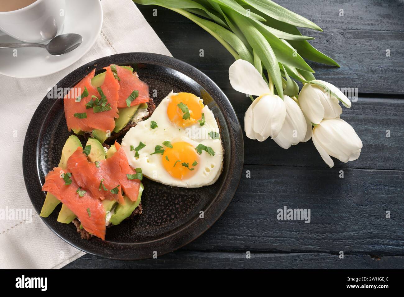 Oeufs frits en forme de coeur et andwichs avec avocat et saumon fumé sur une assiette foncée, serviette blanche et tulipes, bois bleu foncé Banque D'Images