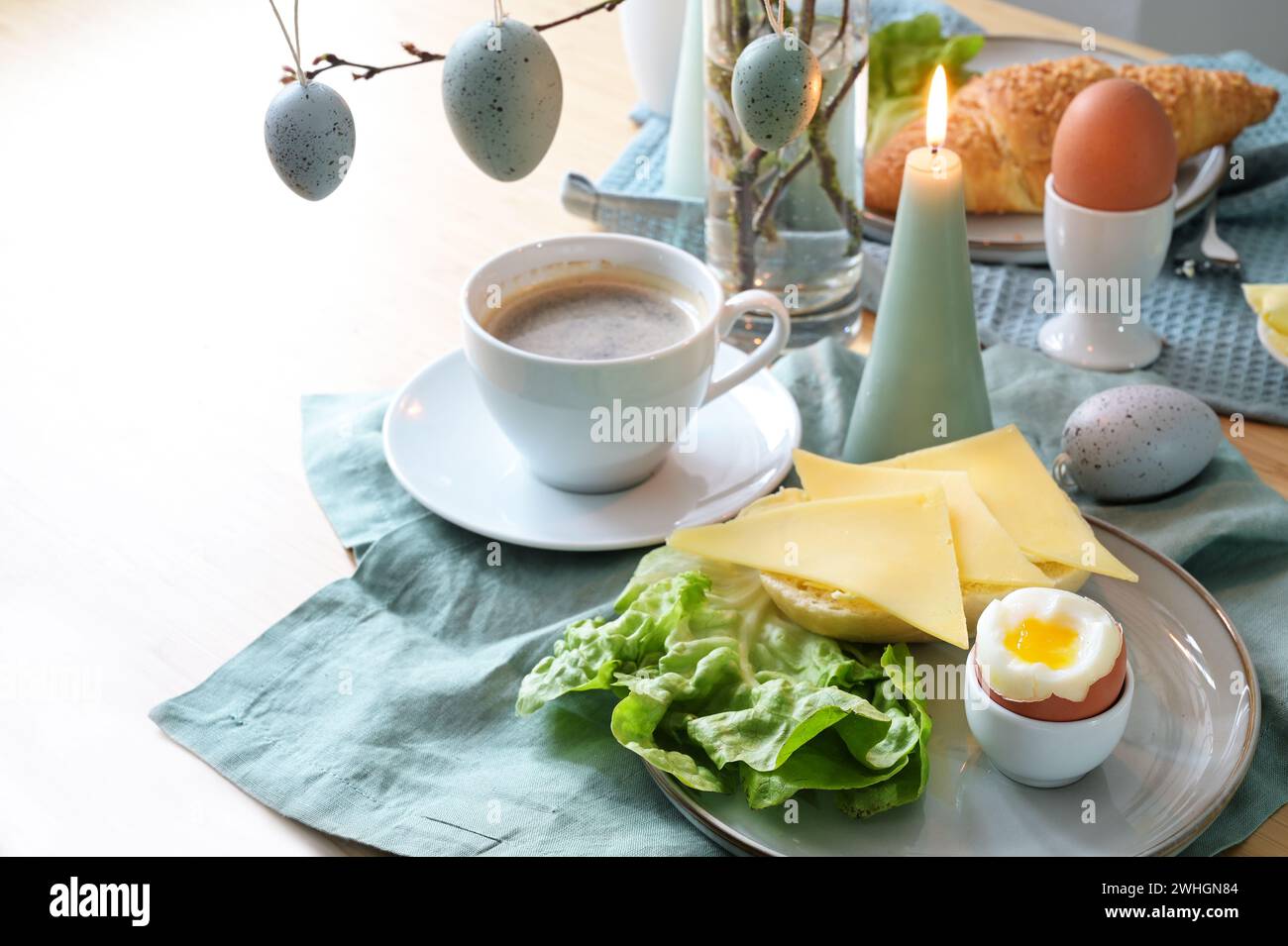 Table de petit déjeuner avec pain, fromage, œuf dur et café sur une serviette turquoise pastel, œufs de Pâques suspendus et canette Banque D'Images