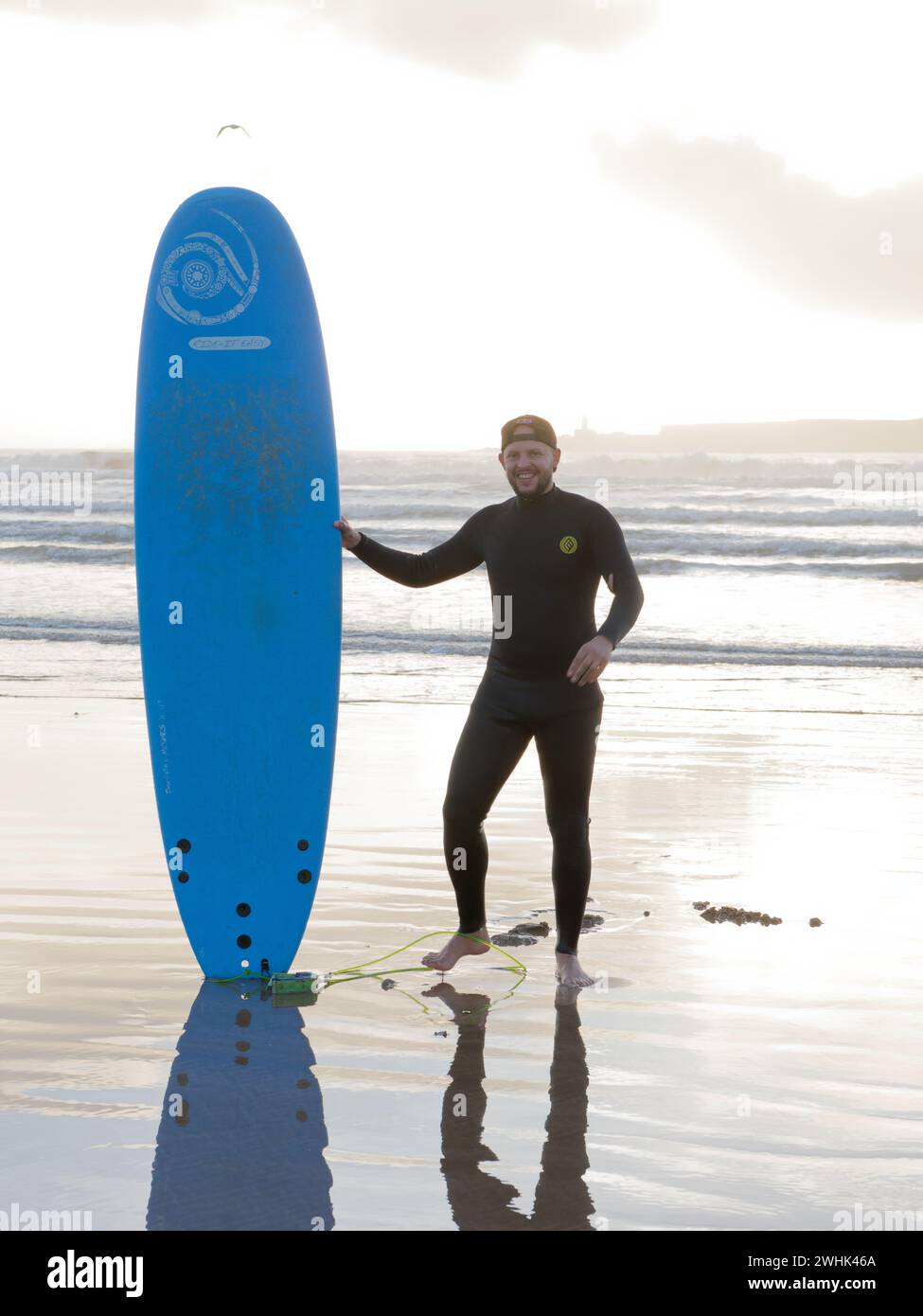 Surfer se tient avec sa planche de surf dans la mer au coucher du soleil avec une île derrière à Essaouira, Maroc. 10 février 2024 Banque D'Images
