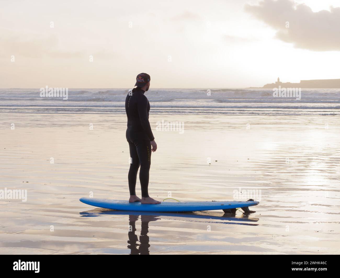 Surfer se tient avec sa planche de surf dans la mer au coucher du soleil avec une île derrière à Essaouira, Maroc. 10 février 2024 Banque D'Images