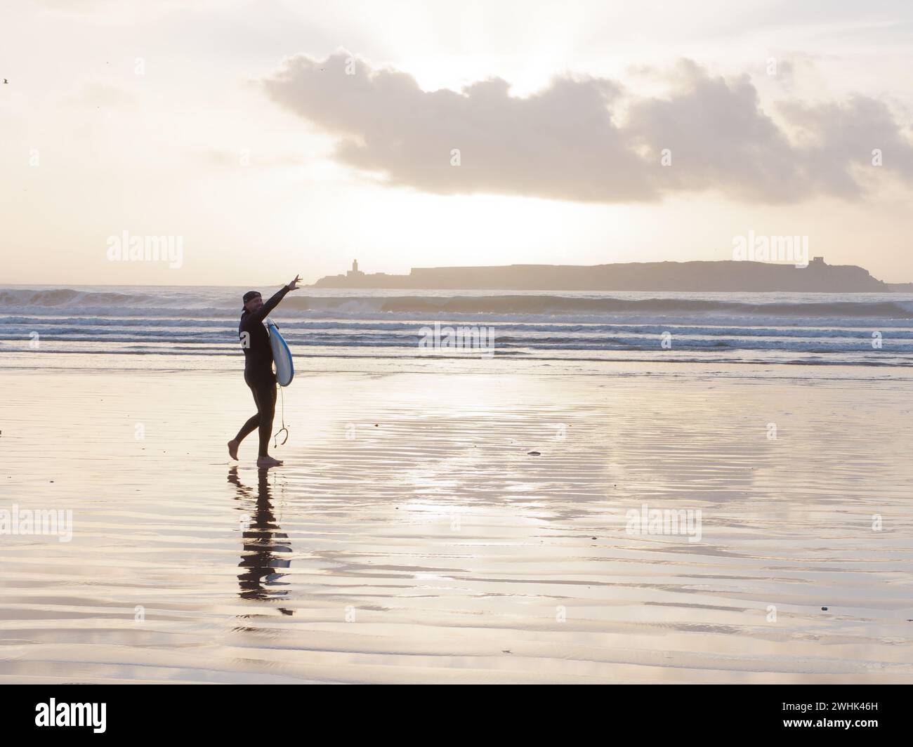 Surfer se tient avec sa planche de surf dans la mer au coucher du soleil avec une île derrière à Essaouira, Maroc. 10 février 2024 Banque D'Images