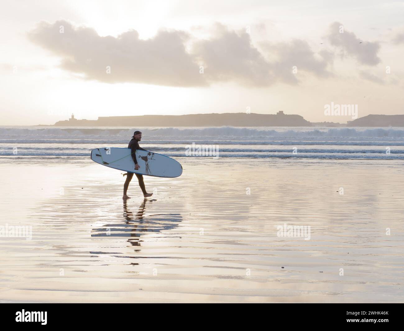 Surfeur se promène avec sa planche de surf dans la mer au coucher du soleil avec une île derrière à Essaouira, Maroc. 10 février 2024 Banque D'Images