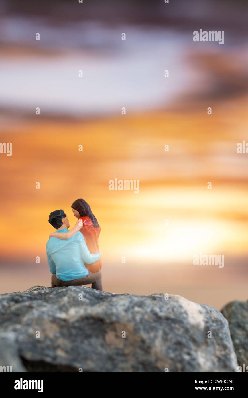 Personnages miniatures, couple assis sur une plage de mer avec fond de coucher de soleil Banque D'Images