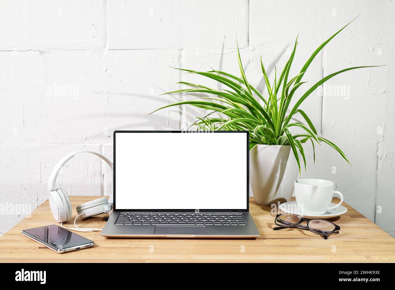 Bureau en bois avec maquette d'ordinateur portable ouverte, écouteurs, téléphone, lunettes et plante devant un mur peint en blanc, bureau, baguette d'affaires Banque D'Images