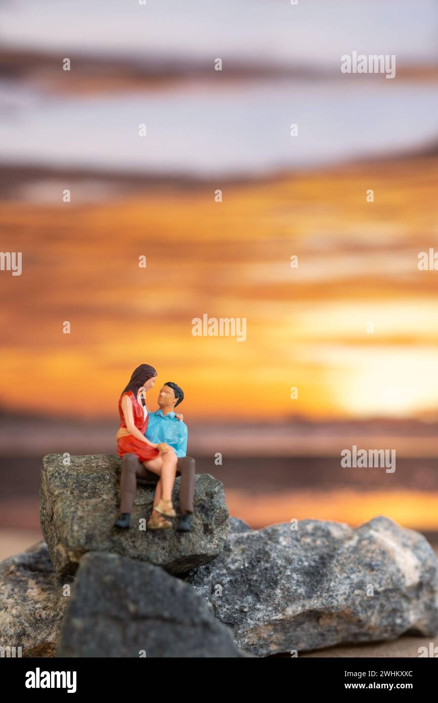 Personnages miniatures, couple assis sur une plage de mer avec fond de coucher de soleil Banque D'Images