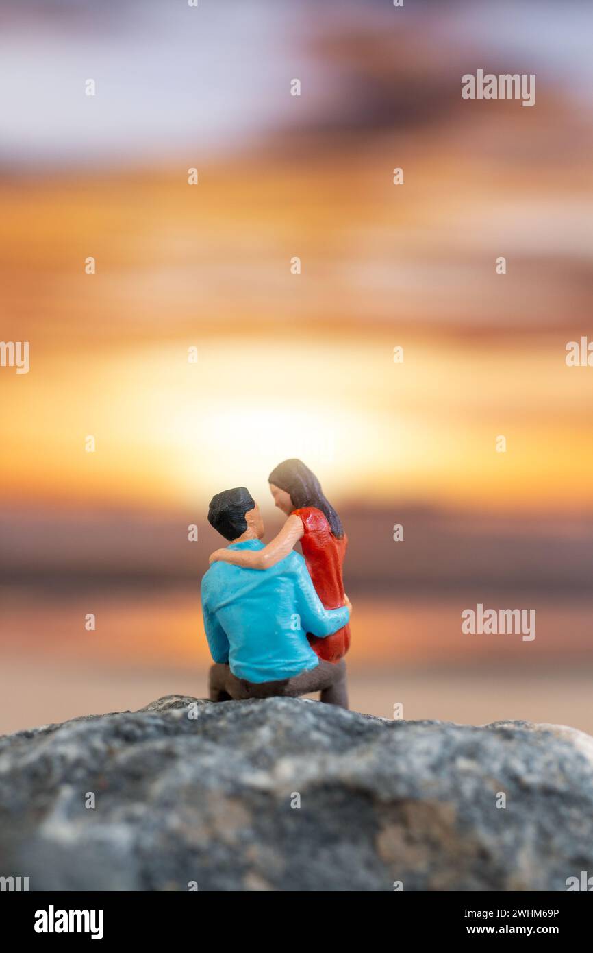 Personnages miniatures, couple assis sur une plage de mer avec fond de coucher de soleil Banque D'Images