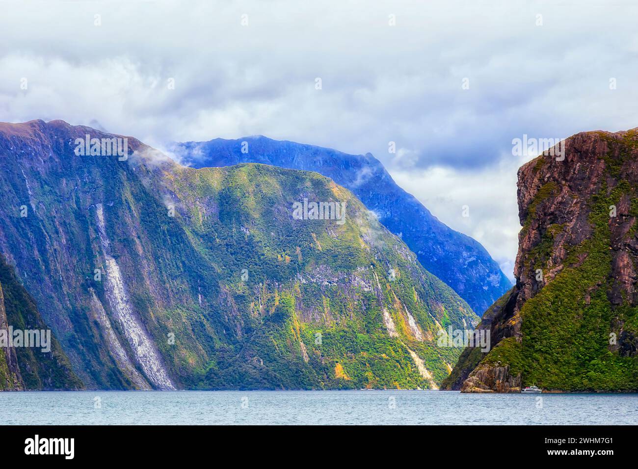 Petit bateau de croisière touristique sous les pics rocheux massifs de Milford Sound dans le fiordland de Nouvelle-Zélande. Banque D'Images