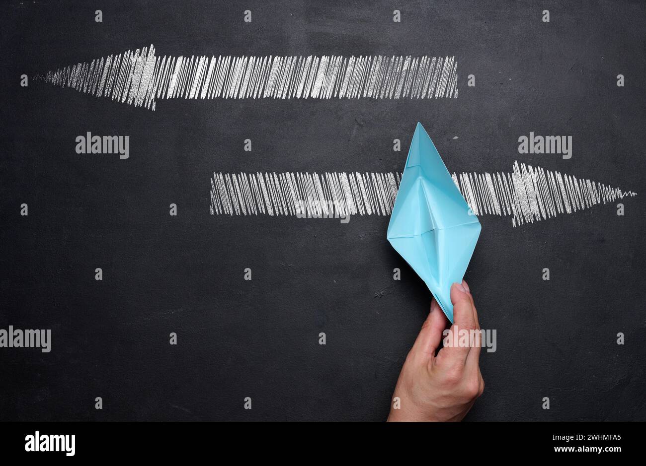 La main d'une femme tient un bateau de papier bleu sur un tableau de craie noire et des flèches dans différentes directions, le choix de la direction de Banque D'Images