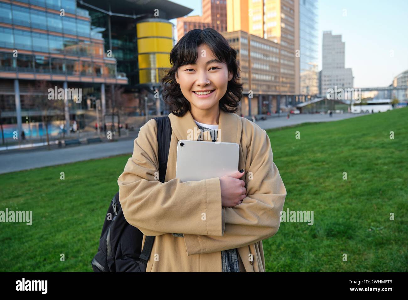 Une fille asiatique heureuse se tient dans la rue, un étudiant de l'université marche avec une tablette numérique dans les mains et les sourires, se tient dans le centre-ville Banque D'Images