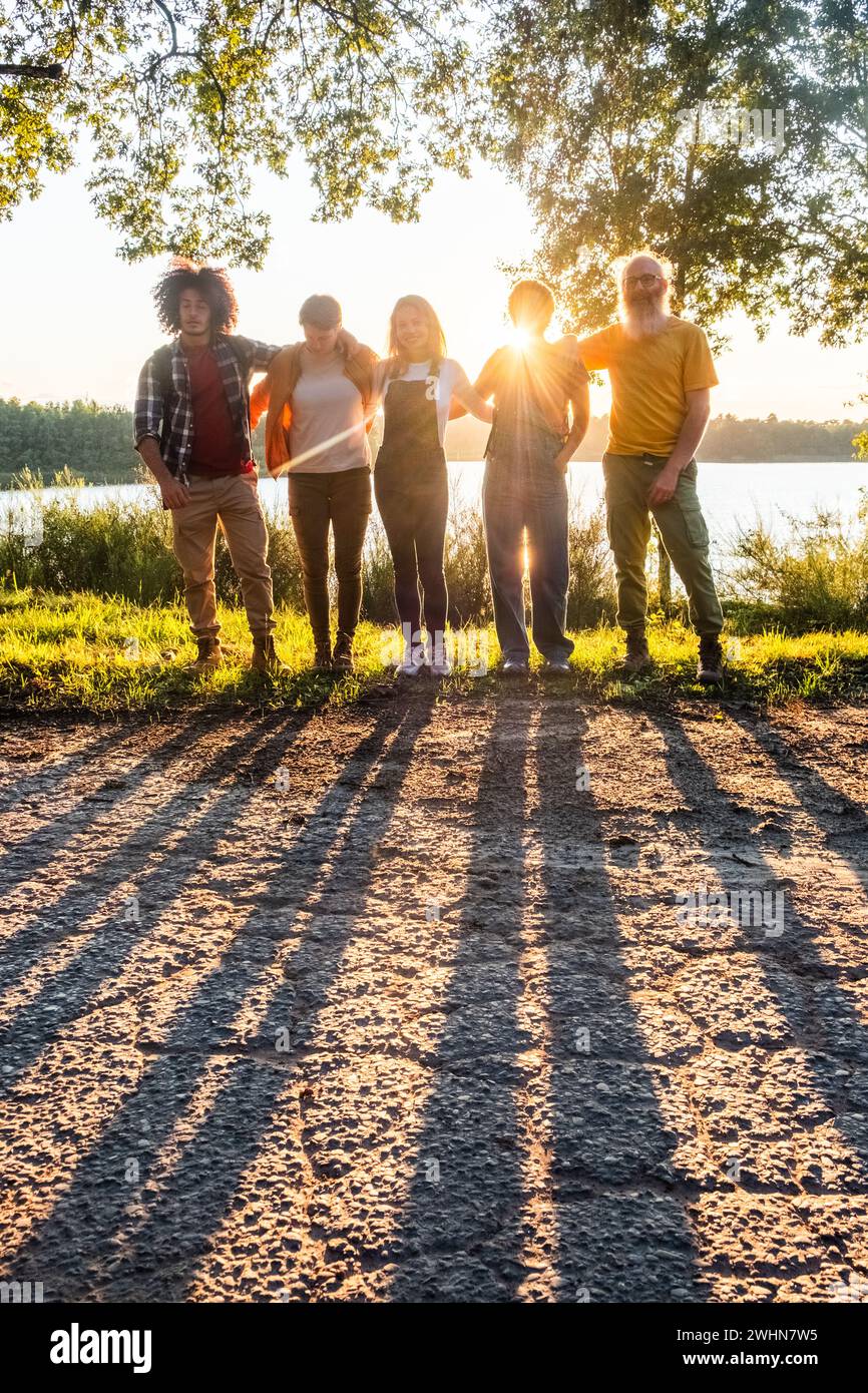 Les ombres de jeunes amis multiraciaux admirant le coucher de soleil doré et s'amusant au bord du lac forestier. Concept de style de vie Banque D'Images