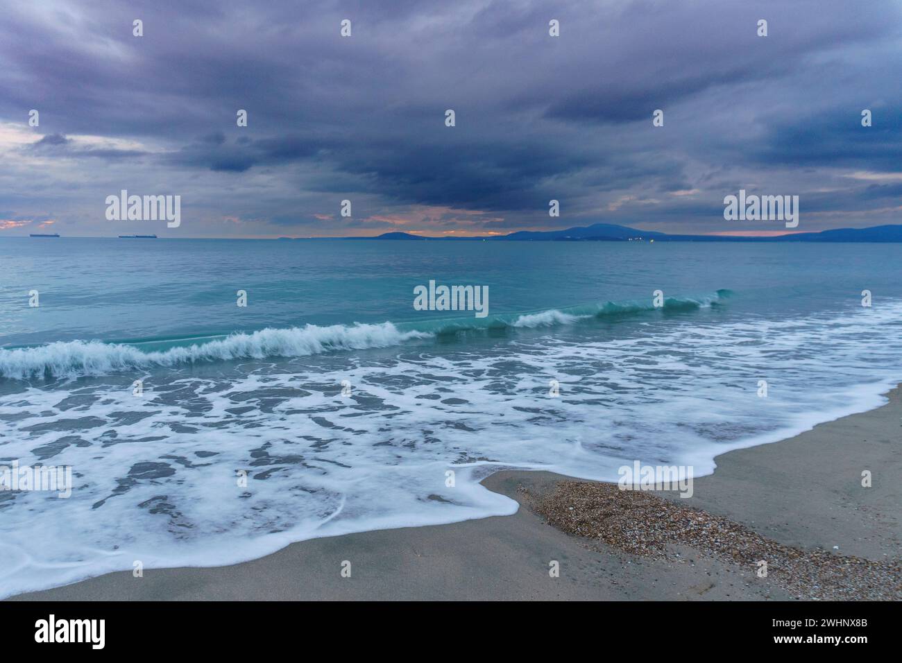 Vagues de l'océan se brisant sur une plage de sable au coucher du soleil, entourées de nuages gris Banque D'Images