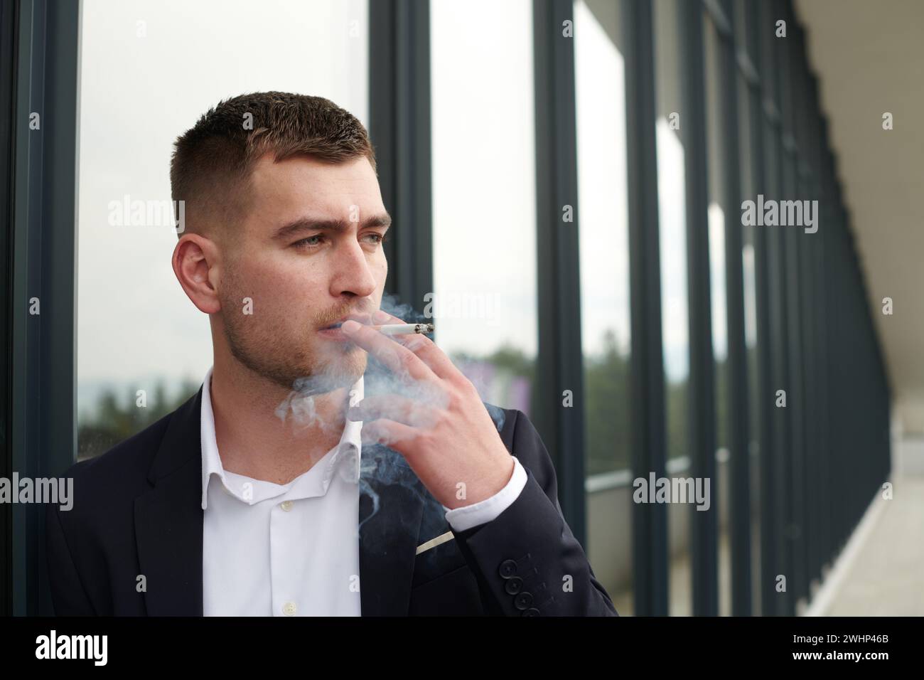 Au milieu de l'agitation de l'entreprise, un homme d'affaires moderne en costume noir prend une pause fumée à l'extérieur de son lieu de travail, à la recherche d'un moment de Banque D'Images