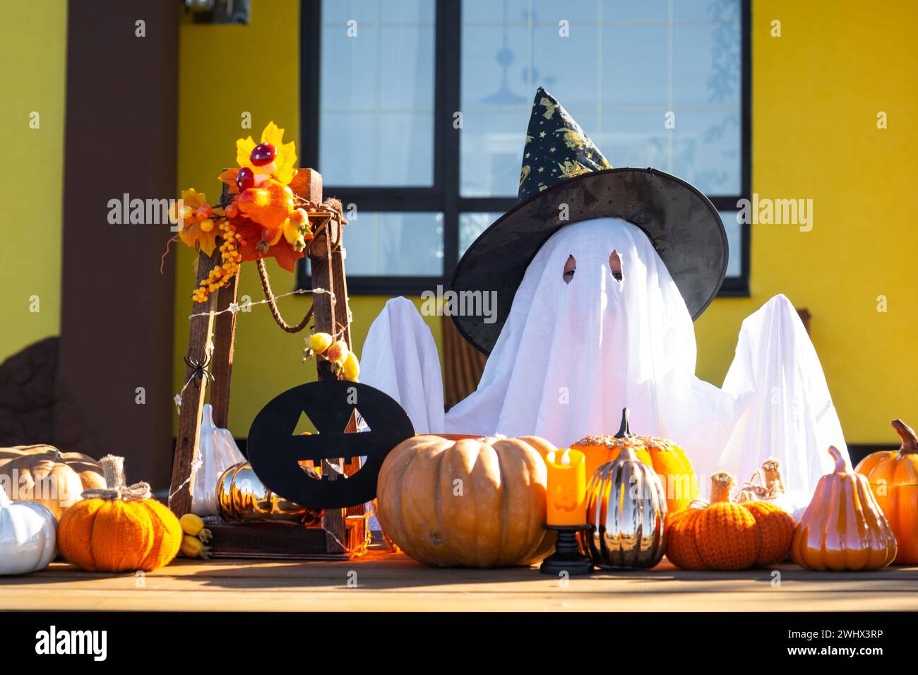Enfant dans le drap de lit avec des fentes comme fantôme dans le chapeau de sorcière et le décor d'Halloween sur le porche de la maison à l'extérieur dans la cour de pum Banque D'Images