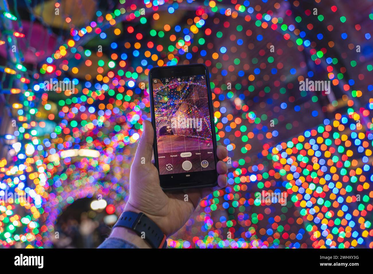 Main masculine tenant un smartphone et prenant une photo avec des lumières de Noël bokeh colorées en arrière-plan Banque D'Images